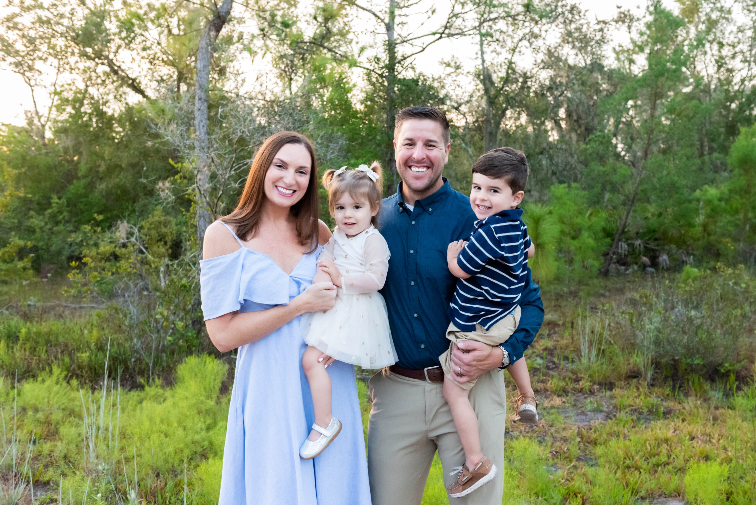 Family of four smiling during a family photography session in a natural outdoor setting in Tampa, Florida, with the parents holding their young son and daughter.