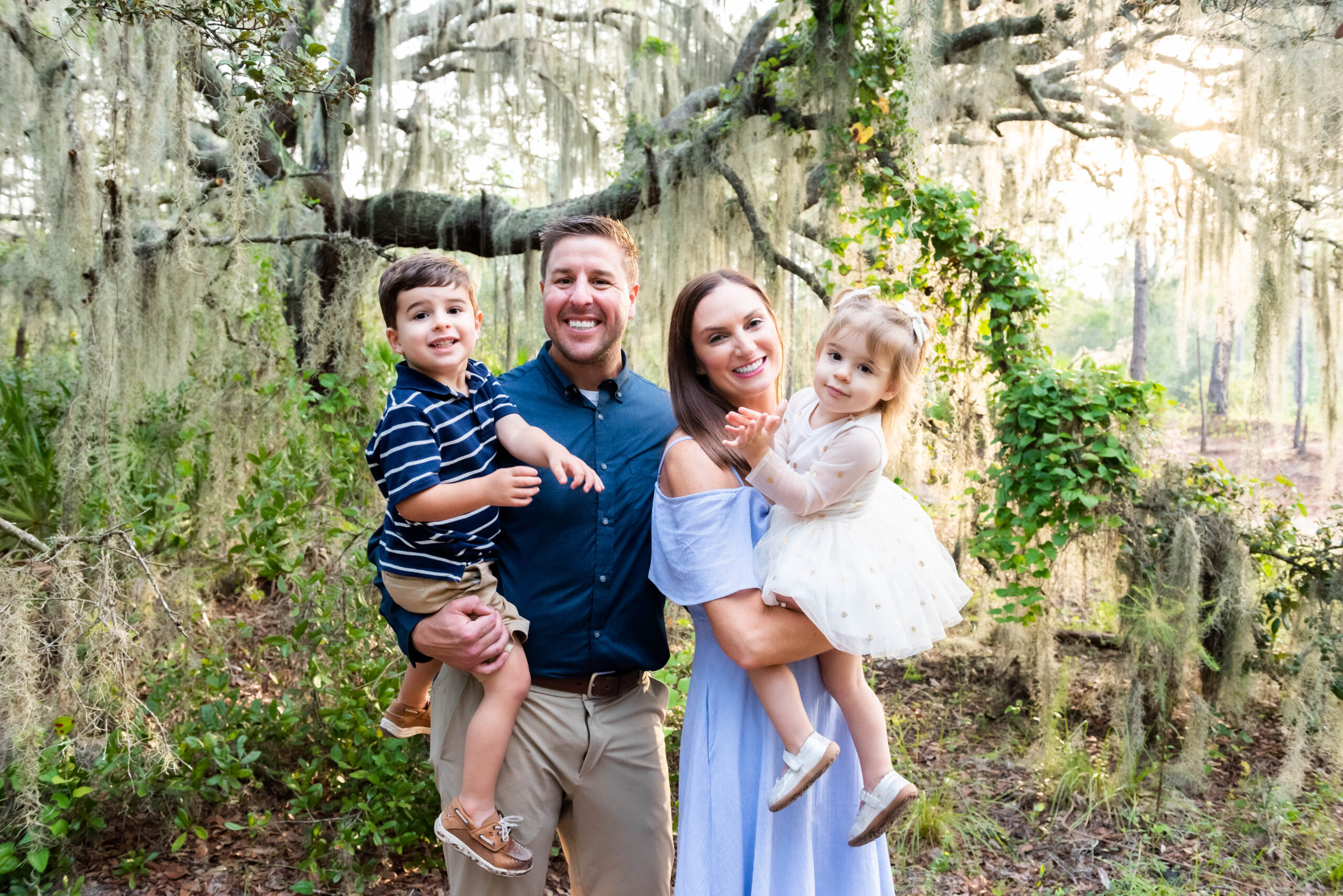 Family of four enjoying a photoshoot in a lush, tree-filled outdoor setting in Tampa, Florida, with the parents holding their young son and daughter.