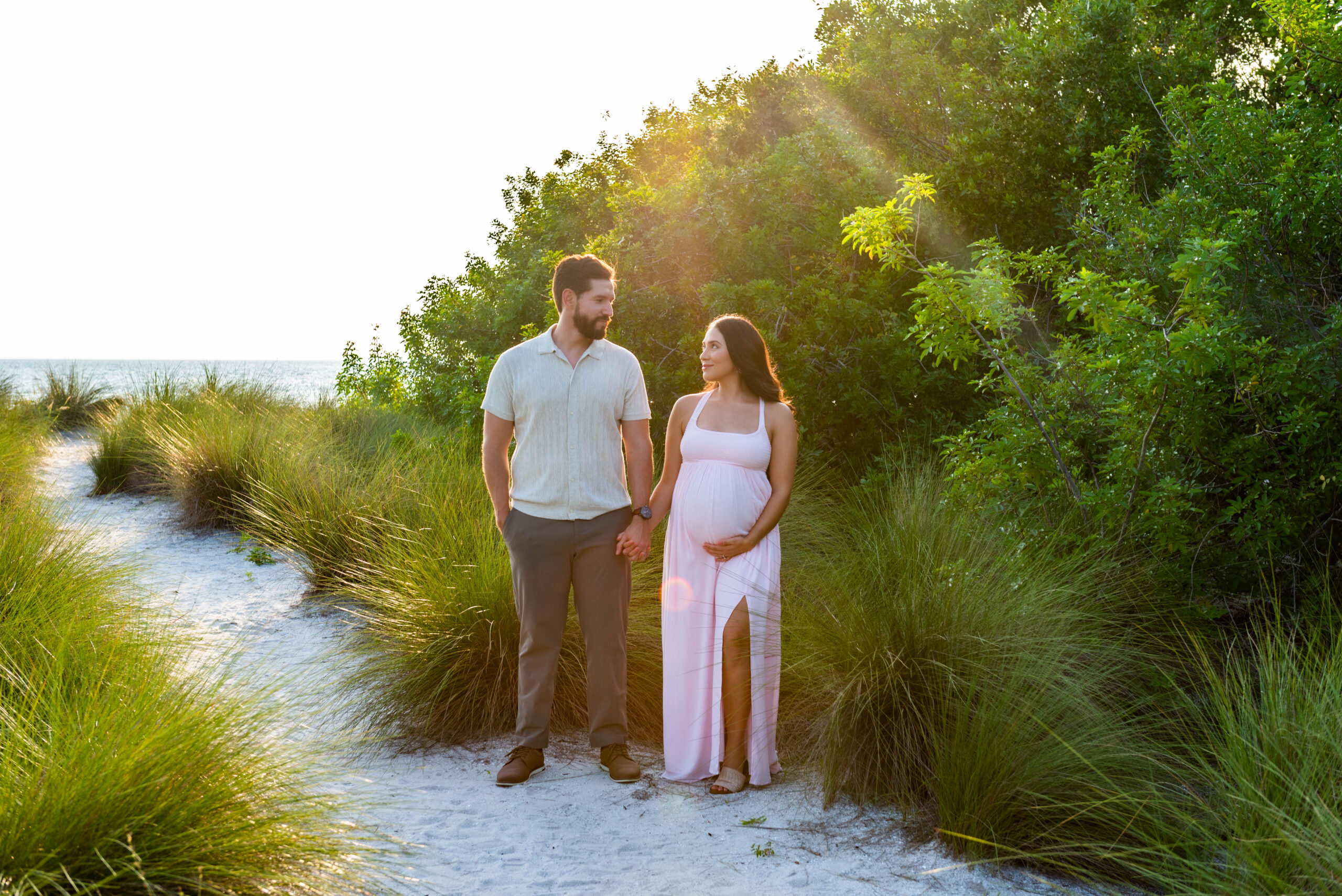 A couple holding hands during a maternity photoshoot at Cypress Point Park, standing on a sandy path surrounded by greenery, with sunlight filtering through the trees.